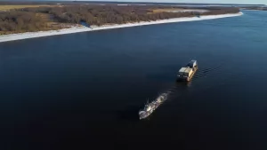 Vista aérea de transporte fluvial em uma barcaça de carga sendo rebocada por um pequeno barco em um grande rio, com margens florestadas e a luz do entardecer refletindo sobre a água.