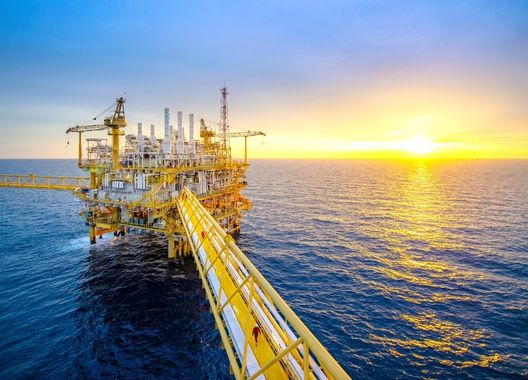 Fotografia de Uma plataforma amarela de petróleo e gás anexada à ponte no meio do mar calmo com céu azul, representando uma empresa offshore