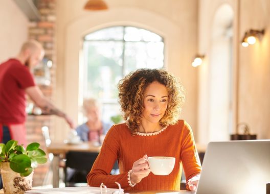 Uma mulher de cabelhos cacheados curtos, segurando uma xícara de café enquanto faz a renovação do seu seguro pelo computador