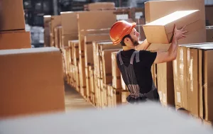 Trabalhador de capacete laranja manuseia caixas em armazenagem logística.