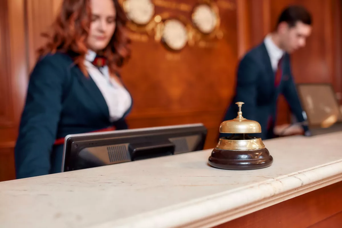 Recepcionistas de hotel em uniformes elegantes ao fundo com balcão de mármore em primeiro plano com campainha de mesa.