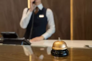 Recepcionista de setor hoteleiro atendendo uma ligação com campainha de mesa em primeiro plano no balcão de madeira.
