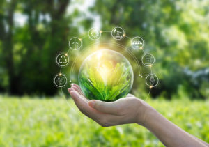 Uma mão segurando um globo transparente com uma planta verde rodeado por ícones relacionados a ecologia e sustentabilidade com um fundo relacionado a natureza. Conceito ecológico sobre impactos ambientais.