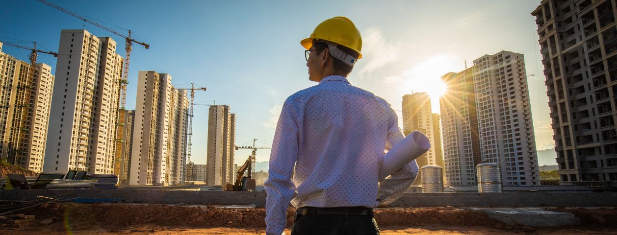 Uma fotografia mostrando engenheiro de costas com planta da obra embaixo do braço e observando o canteiro de obras na construção de prédios a sua frente