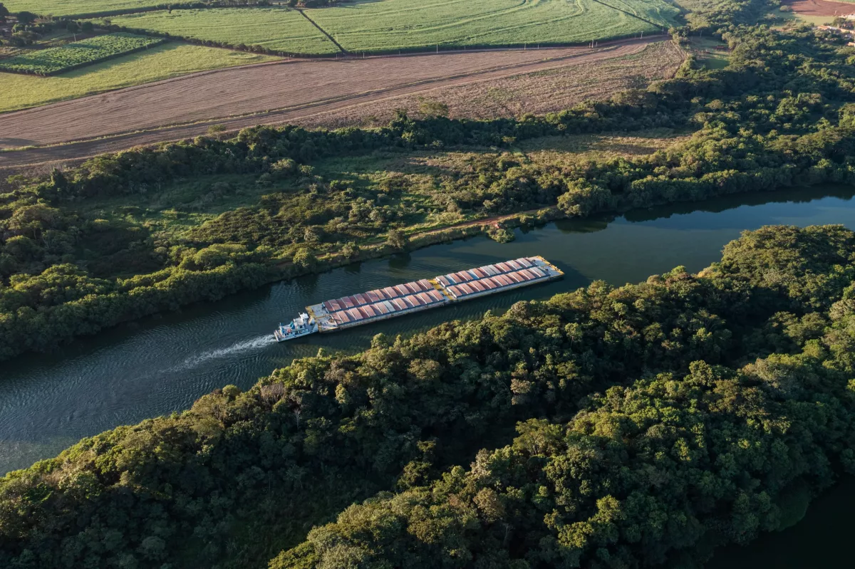 Vista aérea de uma barcaça transportando contêineres de carga através de um rio cercado por uma paisagem diversificada, com áreas de mata nativa e campos agrícolas ao entardecer.