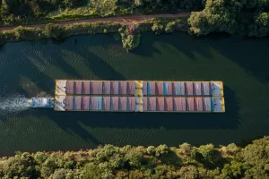 Foto aérea de transplante fluvial em uma barcaça de carga carregada com diversos contêineres atravessando um rio serpenteante, cercado por vegetação densa, em um dia ensolarado.