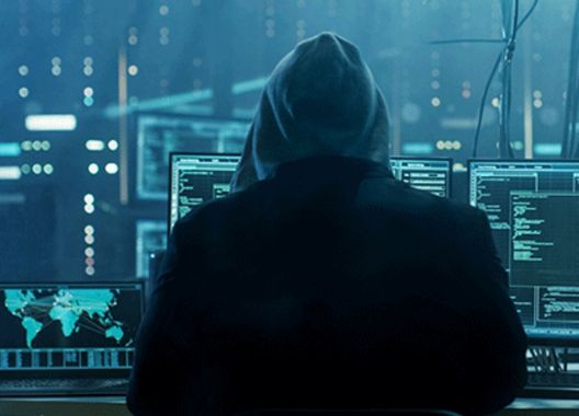 uma foto de uma hacker emcapuzado olhando computadores sincronizados em um ataque hacker