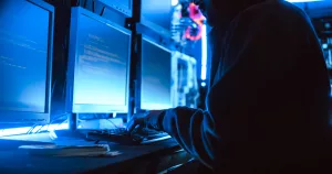 Hacker com múltiplos monitores com códigos de programação, em um ambiente de servidor, enfatizando os ataques cibernéticos no Brasil.