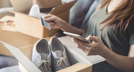 Uma fotografia de mulher com uma caixa aberta em seu colo com um tênis dentro recém comprado na black friday e com um cartão de crédito e um celular nas mãos fazendo compras online