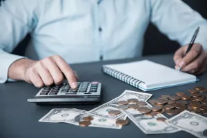 Homem calculando finanças com calculadora, dinheiro e moedas sobre a mesa, simbolizando as possíveis causas de calote financeiro