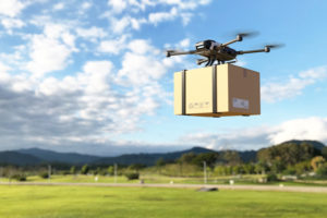 drone prata 4 hélices transportando uma encomenda com uma ambientação do campo ao fundo e o céu azul