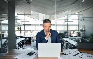 Homem de empresas com terno em escritório olhando para o notebook com parede de vidro transparente ao fundo