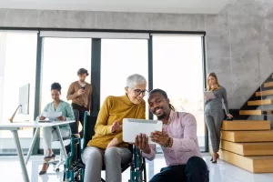 homem jovem agachado ensinando mulher idosa em cadeira de rodas a utilizar um tablet em ambiente corporativo ao fundo representando a inclusão no mercado de trabalho