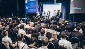 Vista traseira de um público diversificado em um evento corporativo, com foco em vários profissionais sentados em cadeiras, voltados para um painel de palestrantes no palco iluminado.