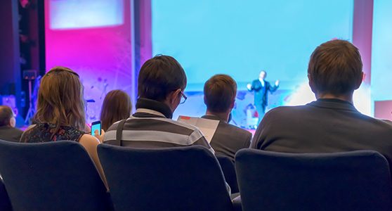 Uma fotografia de uma plateia assistindo a um homem palestrando em um palco em evento