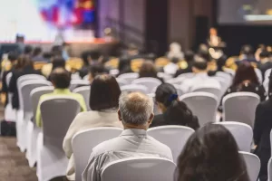 Pessoas sentadas em cadeiras brancas assistindo a uma apresentação em um auditório, foco no homem de cabelos grisalhos no centro.