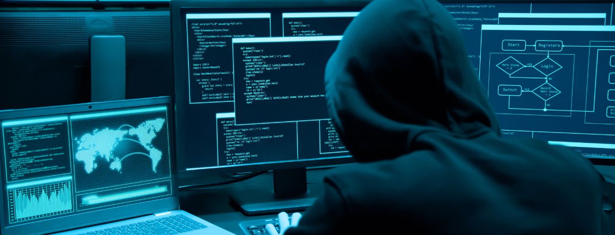 Uma fotografia de mostrando um jovem hacker sentado e invadindo três computadores ao mesmo tempo através de Ransomware