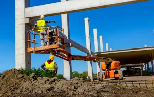 um canteiro de obras onde um trabalhador em uma plataforma elevatória está realizando trabalhos em colunas de concreto. Um outro trabalhador, usando um colete refletor e capacete, está no chão perto de um monte de terra. No fundo, pode-se ver maquinário pesado e a estrutura parcialmente construída de um edifício ou ponte.