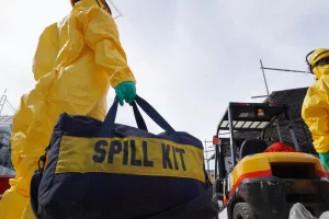 Um trabalhador em traje de proteção amarelo carrega um kit de contenção de derramamento, preparando-se para responder a um vazamento de produtos químicos.