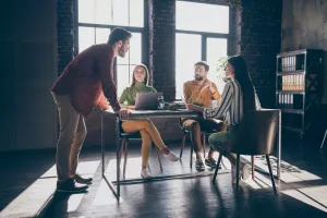 Funcionários de empresa de pequeno porte reunidos em sala de reunião com três pessoas sentadas e uma pessoa de pé apoiada na mesa