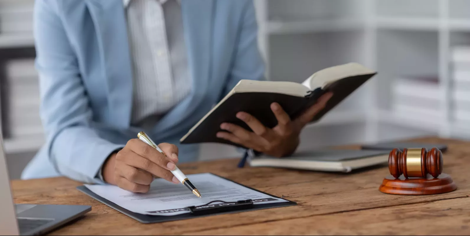 advogada segurando um livro com a mão esquerda e fazendo anotações com a mão direita usando paletó com um martelo ao lado