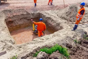 trabalhadores em canteiro de obras com um trabalhador de laranja dentro de buraco no chão de terra enquanto outro trabalhador está na parte superior do chão.