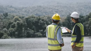 dois engenheiros ambientais com colete e capacete de construção olhando para plantas de obras em frente de floresta