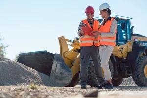 dois engenheiros com coletes de proteção laranja conversando sorrindo sobre projeto com escavadeira amarela ao fundo
