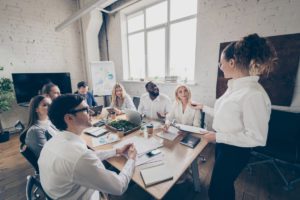 Sala de reunião com várias pessoas sentadas em volta de uma mesa e uma mulher de pé fazendo uma apresentação em ambiente empresarial.