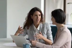 Duas mulheres conversando em um escritório com roupas formais em a frente de uma mesa com um notebook