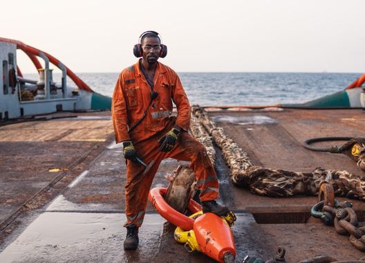 Uma fotografia de um funcionário de empresa offshore em uma plataforma em alto mar, sujo de petróleo com o pé sob uma âncora de navio